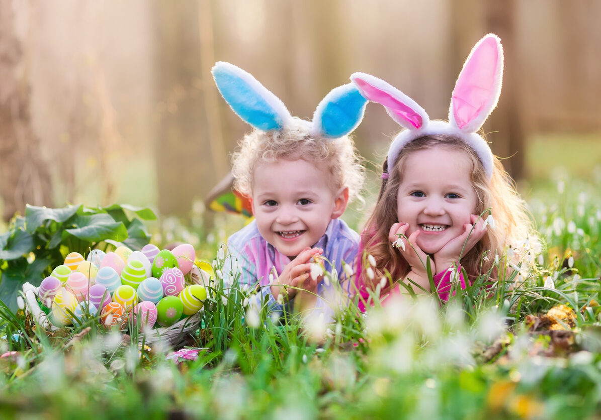 Kids on Easter egg hunt in blooming spring garden. Children with bunny ears searching for colorful eggs in snow drop flower meadow. Toddler boy and preschooler girl in rabbit costume play outdoors.